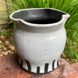 May include: A ceramic pitcher with a wide, rounded body and a flared, wavy rim. The exterior features a white crackle glaze finish, while the interior is dark gray. The base is decorated with a series of vertical black stripes around the bottom edge.