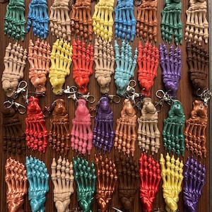 Puede incluir: Una colección de llaveros de mano de esqueleto impresos en 3D y coloridos. Los llaveros son de varios colores, incluyendo verde, azul, naranja, amarillo, rojo y morado. Cada llavero tiene un cierre plateado.