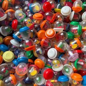 May include: A collection of colorful plastic toy capsules. The capsules are clear with various colored lids and contain small toys or figures. The lids are in shades of red, orange, blue, and white. Some capsules have images of fruits and animals.