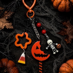 Op de afbeelding: Een sleutelhanger met een oranje hartvormige sluiting, een stervormige bedel met een candy corn hanger, een maansikkel pompoen lolly bedel en een skeletfiguurtje met rode en zwarte kralen aan een zilverkleurige metalen ketting.