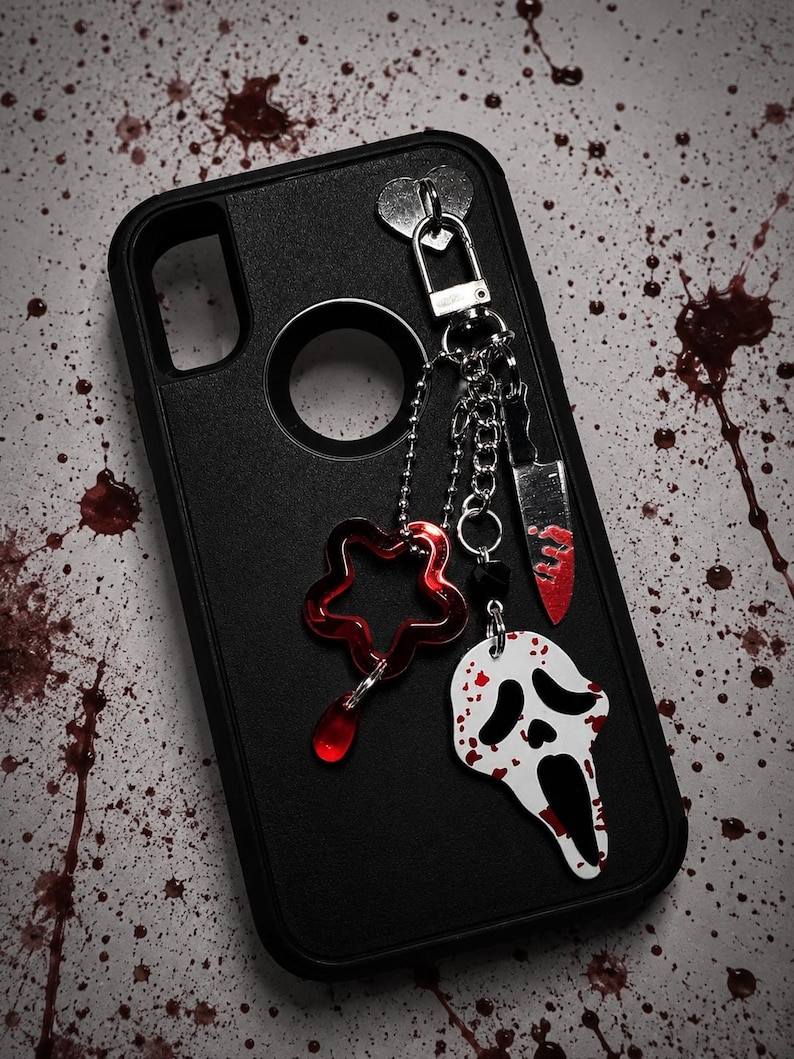 May include: Black phone case with a silver-tone metal clasp and dangling charms. Charms include a red star, a white and black ghost face, and a knife with red accents. The background has red splatter.