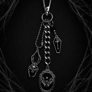 May include: A silver-toned metal keychain featuring a lobster clasp and a thick chain. Attached are three charms: a spider with a skull-shaped body, a small coffin charm with a cross design, and an oval frame pendant containing a bat silhouette.
