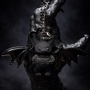 May include: A black bat keychain with a plush body and shiny, detailed wings. The bat has a lace collar and is attached to a silver chain and key ring. The background is dark and moody.