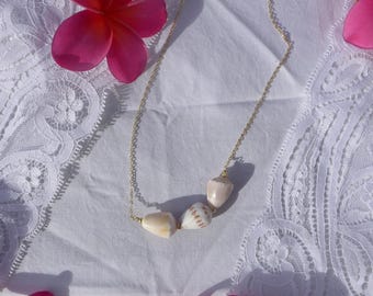 May include: A delicate gold chain necklace featuring four natural seashells. The shells are cream-colored with subtle patterns, strung along a fine gold chain. Pink flowers are scattered around the necklace on a white lace background.