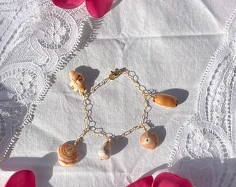 May include: A gold-toned chain bracelet adorned with various seashells. The bracelet features a delicate chain design and is embellished with several uniquely shaped shells in shades of beige and brown. Pink flowers are scattered around the bracelet.