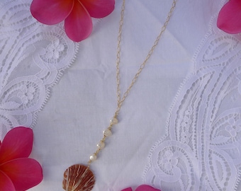 May include: A gold-colored necklace with a seashell pendant and small pearls. The seashell is brown and white, and the necklace is set against a white lace background with pink flowers.
