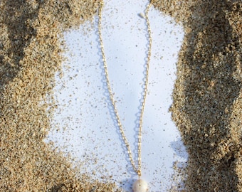 May include: A delicate gold chain necklace with a single pearl pendant. The necklace is displayed on a white surface against a backdrop of sand.
