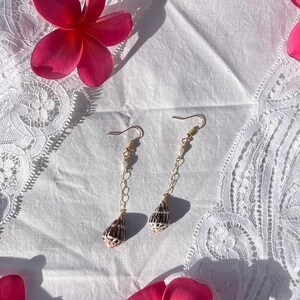 Puede incluir: Un par de pendientes dorados con diseño de cadena, pequeñas cuentas blancas y colgantes de concha. Los pendientes se exhiben sobre una superficie de encaje blanco, con flores rosas en el fondo.