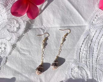May include: A pair of gold-toned earrings with a chain design, featuring small white beads and seashell pendants. The earrings are displayed on a white lace surface, with pink flowers in the background.
