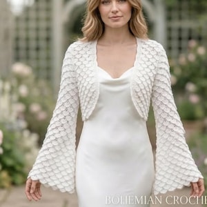 May include: A white, crochet wedding shrug with flared sleeves. The shrug is worn over a white dress. The image is set outdoors with a garden background. Text at the bottom reads "Bohemian Crochet Wedding Shrug".