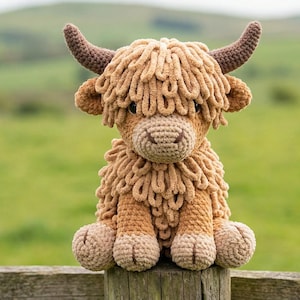 May include: A crocheted Highland cow toy, featuring a light brown body, darker brown horns, and a shaggy mane. The toy is positioned on a wooden fence post, set against a blurred green backdrop.