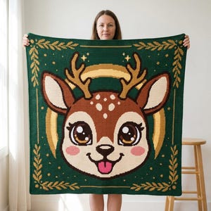 May include: A square crochet blanket in dark green featuring a central graphic of a brown deer head with large eyes, pink cheeks, and gold antlers. The design is framed by a gold leaf border with small stars, creating a detailed tapestry-style pattern.
