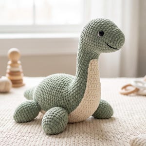 Op de afbeelding: Een gehaakt dinosaurusspeeltje met een saliegroen lichaam en een crèmekleurige borst. Het pluche wezen heeft een lange nek, een ronde kop met zwarte knoopogen, een gestikte glimlach en vier korte, dikke pootjes met een geknoopte textuur.