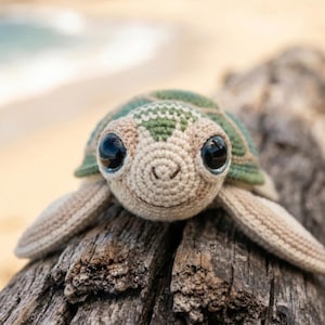 May include: A crocheted sea turtle with a green and beige shell, large black eyes, and a smiling face. The handmade turtle is lying on a piece of weathered wood, with a blurred beach and ocean in the background.