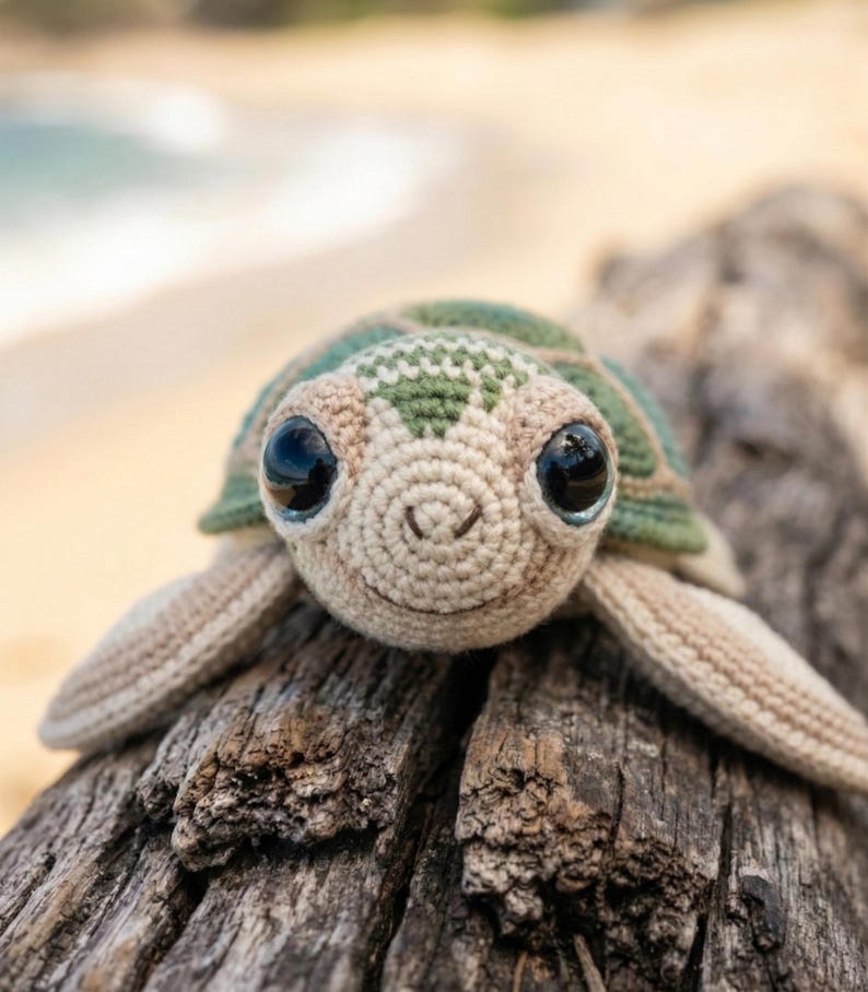 May include: A crocheted sea turtle with a green and beige shell, large black eyes, and a smiling face. The handmade turtle is lying on a piece of weathered wood, with a blurred beach and ocean in the background.