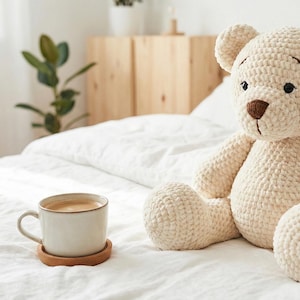 May include: A cream-colored, crocheted teddy bear with a brown nose and black eyes sits on a white bed. A white mug of coffee rests on a wooden coaster. The scene is set in a bright, airy bedroom.
