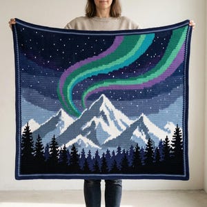 May include: A square crochet blanket featuring a nighttime mountain landscape. The design displays a dark blue sky with white stars, a vibrant green and purple aurora borealis, snow-capped mountain peaks, and a dense forest of black pine trees.