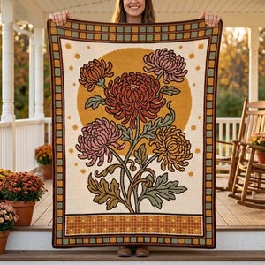 Op de afbeelding: Een rechthoekige gehaakte deken met een grote centrale afbeelding van roestkleurige, roze en gouden chrysanten tegen een gele zonachtergrond. De rand heeft een ruitpatroon in bruin, groen en goud met kleine steraccenten.