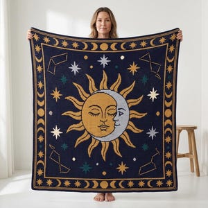 May include: A square woven blanket featuring a dark navy blue background with a central golden sun and silver crescent moon face. The border displays a repeating pattern of moon phases and stars, with additional constellations and stars scattered.