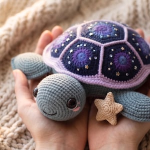 May include: A handcrafted crochet turtle toy. The turtle has a blue and purple shell with a starry night design, a light purple border, and gold star embellishments. A small, tan starfish is also present.