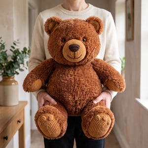 Könnte beinhalten: Ein großer, plüschiger brauner Teddybär mit weicher, flauschiger Textur. Der Bär hat eine gehäkelte Schnauze in einem helleren Beigeton, eine dunkelbraune gestickte Nase sowie passende gehäkelte Details an den Innenohren und den Fußsohlen.