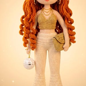 May include: Crocheted doll in a gold and cream outfit, holding a disco ball and a gold clutch. The doll has long, curly red hair, gold hoop earrings, and a gold necklace. The doll is wearing a gold top, cream trousers, and gold platform sandals.