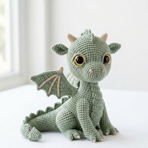 May include: A crocheted dragon in shades of green, with large, golden-yellow eyes. The handmade dragon has small wings, horns, and a long tail. The dragon is sitting on a white surface.