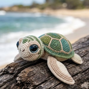 Könnte beinhalten: Eine handgefertigte Häkelschildkröte mit grün-beigem Panzer und großen, dunklen Augen. Die Schildkröte liegt auf einem Stück Treibholz, mit einem verschwommenen Strand und Ozean im Hintergrund. Der Panzer der Schildkröte hat ein geometrisches Muster.