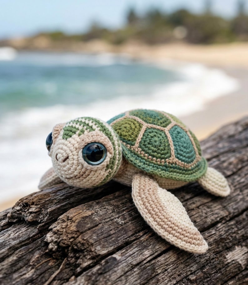 May include: A handmade crochet turtle with a green and beige shell and large, dark eyes. The turtle is resting on a piece of driftwood, with a blurred beach and ocean background. The turtle's shell has a geometric pattern.