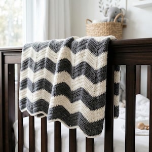Könnte beinhalten: Ein dunkelbraunes Kinderbett mit einer grau-cremefarbenen Decke mit Zickzackmuster, die über die Seite drapiert ist. Die Decke besteht aus einem weichen, strukturierten Material. Ein Weidenkorb mit einem Stoffelefanten ist im Hintergrund zu sehen.