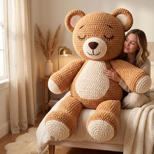 May include: A large, handmade crochet teddy bear in shades of tan and cream. The bear has a round head, closed eyes, and a cream-colored belly and paws. The bear is sitting on a bed.