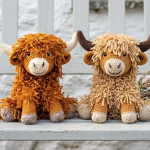 May include: Two handmade crochet highland cow plush toys sitting side by side. The left cow is dark orange with cream horns, while the right cow is light tan with dark brown horns. Both feature shaggy yarn bodies, round black eyes, and stitched mouths.