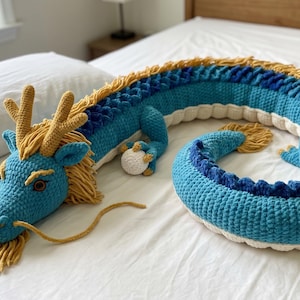 Puede incluir: Un gran dragón de ganchillo hecho a mano en tonos azules, amarillos y blancos. El dragón tiene una cabeza detallada con cuernos y un cuerpo largo. El dragón está sobre una colcha blanca.