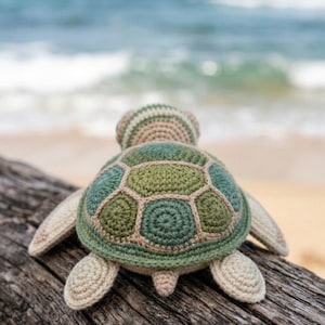 May include: A handmade crochet turtle toy in shades of green, beige, and brown, resting on a weathered wooden log. The turtle's shell features a geometric pattern, and its flippers and head are visible. The background shows a blurred ocean and sandy beach.