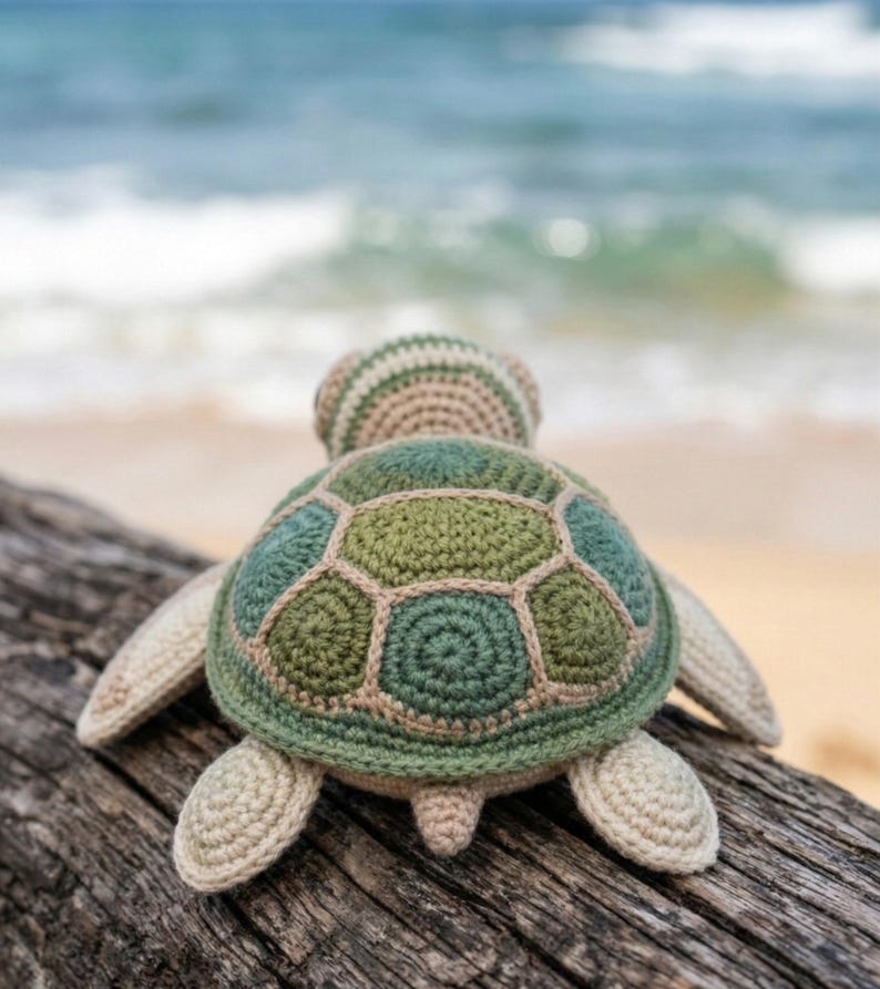 May include: A handmade crochet turtle toy in shades of green, beige, and brown, resting on a weathered wooden log. The turtle's shell features a geometric pattern, and its flippers and head are visible. The background shows a blurred ocean and sandy beach.