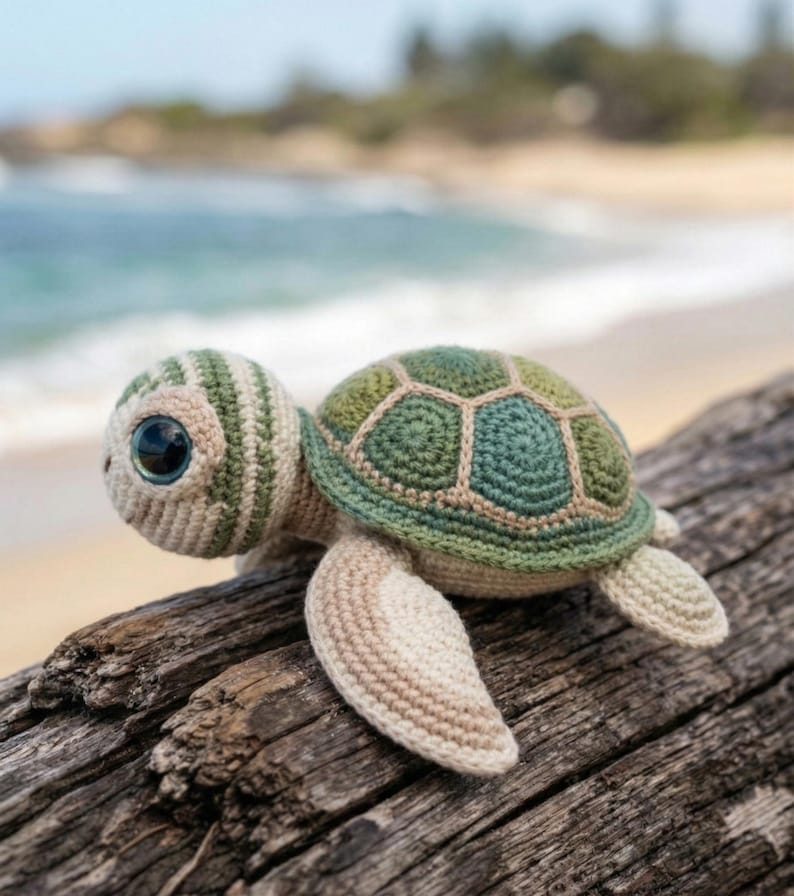 May include: A crocheted sea turtle with large, expressive eyes. The turtle features a green and beige shell, a striped head, and beige flippers. It is resting on a piece of driftwood, with a blurred beach and ocean background.