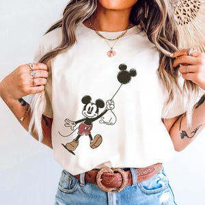 May include: Ivory t-shirt with a Mickey Mouse graphic holding a black balloon with "Smile" on the string. Mickey wears red shorts and brown shoes. The shirt is worn with light blue denim shorts. The word "IVORY" is printed vertically on the left.