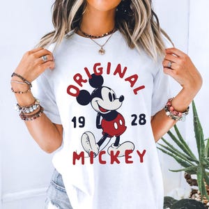 May include: White t-shirt featuring a graphic of Mickey Mouse with the text "ORIGINAL MICKEY 1928" in red and black. The design includes a smiling Mickey Mouse in his classic outfit. The shirt is a casual, everyday wear item.