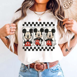 May include: Ivory-colored t-shirt featuring three Mickey Mouse illustrations within a black and white checkered border. Mickey is depicted in various poses, wearing his signature red shorts and white gloves. The shirt is paired with light blue denim shorts and a brown belt.