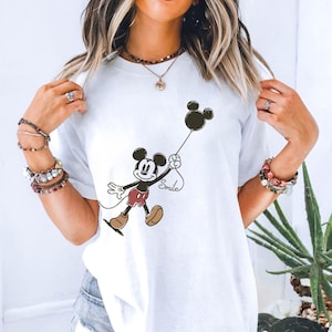 May include: White t-shirt featuring a cartoon Mickey Mouse holding a black balloon with the word "Smile" written below. Mickey is wearing red shorts and brown shoes. The shirt is a casual style.