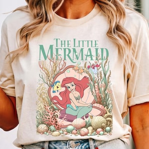 Può includere: T-shirt color avorio con grafica di Ariel de La Sirenetta. Il design include il testo "The Little Mermaid" in verde acqua, con Ariel su una conchiglia, circondata da coralli, pesci e conchiglie. La maglietta è abbinata a pantaloncini di jeans.