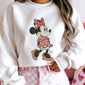 May include: A white sweatshirt featuring a Minnie Mouse graphic. Minnie is depicted in her classic outfit: a red polka-dot dress and bow, and brown shoes. The Disney logo is visible. The model is wearing pink checkered shorts.