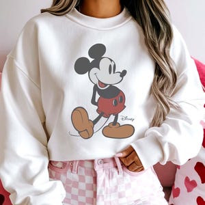 May include: A white sweatshirt featuring a graphic of Mickey Mouse in his classic pose. Mickey is depicted in black, red, and tan colors. The Disney logo is printed on the lower right side of the graphic. The sweatshirt has long sleeves.