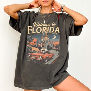 May include: Dark gray t-shirt with a graphic that says "Welcome to Florida" and features a vintage car with Mickey and Minnie Mouse driving past a castle. The shirt has a vintage, worn look.