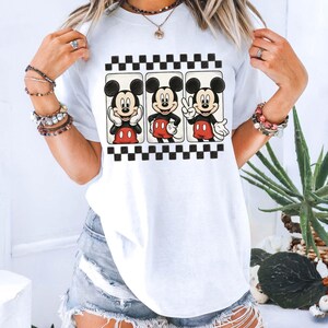 May include: White t-shirt featuring three Mickey Mouse illustrations in black frames with a black and white checkered border. Mickey is depicted in various poses, wearing red shorts and white gloves. The shirt is a casual, everyday wear item.
