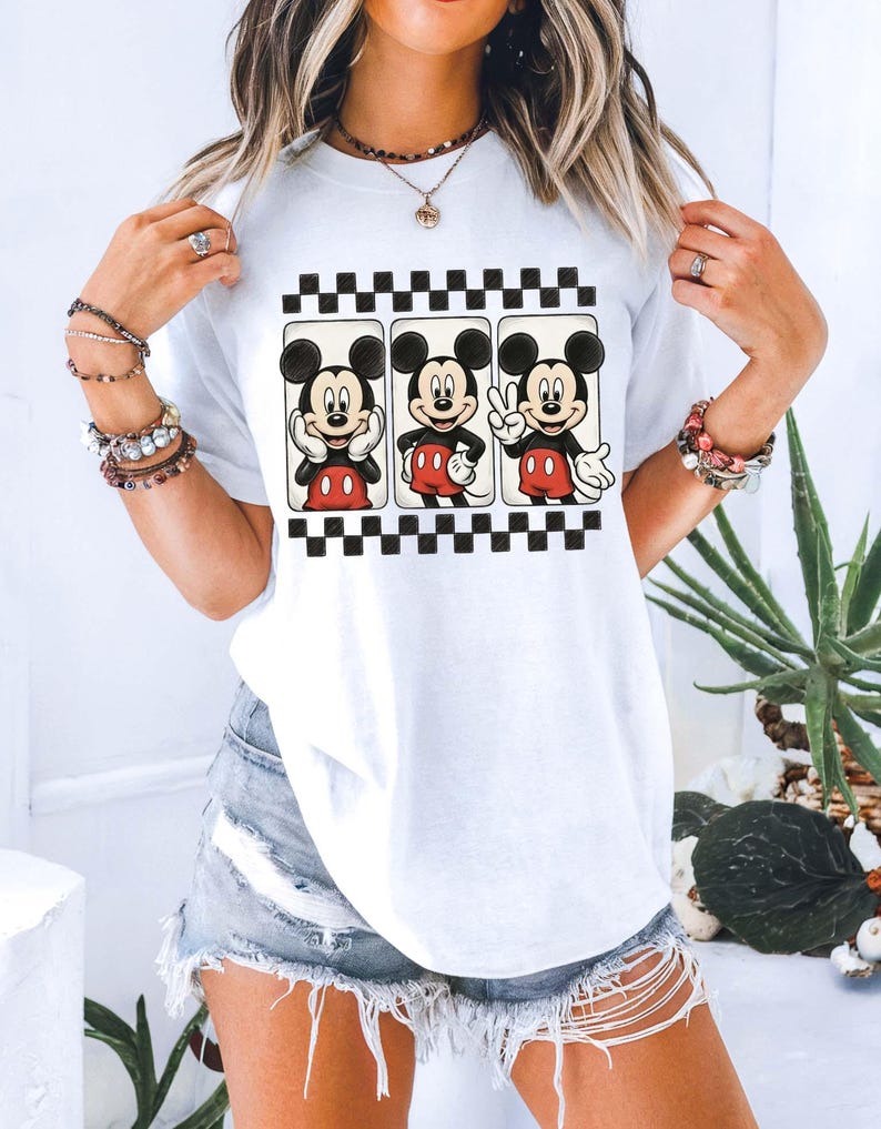 May include: White t-shirt featuring three Mickey Mouse illustrations in black frames with a black and white checkered border. Mickey is depicted in various poses, wearing red shorts and white gloves. The shirt is a casual, everyday wear item.
