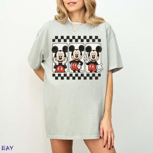 May include: A light green t-shirt featuring a graphic of Mickey Mouse in three different poses. The graphic is framed by a black and white checkered border. Mickey Mouse is wearing red shorts and white gloves.