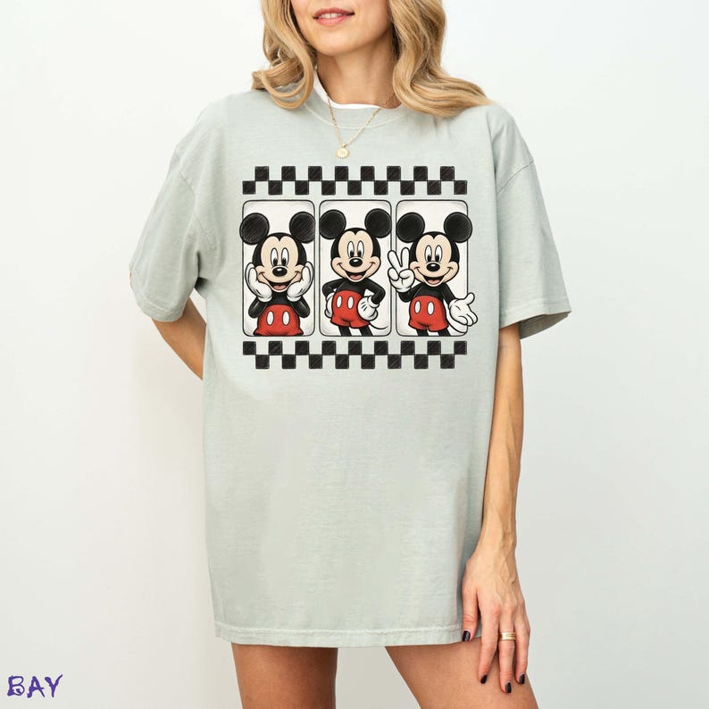 May include: A light green t-shirt featuring a graphic of Mickey Mouse in three different poses. The graphic is framed by a black and white checkered border. Mickey Mouse is wearing red shorts and white gloves.