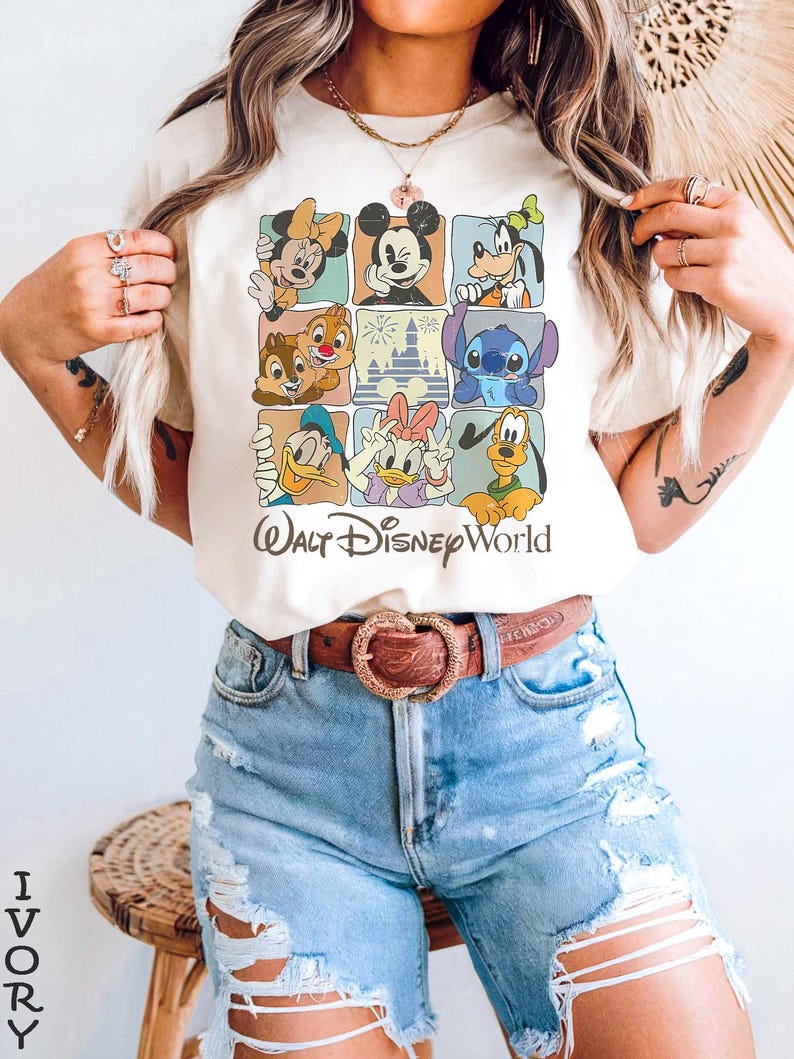 Op de afbeelding: Ivoor T-shirt met een raster van Disney-personages, waaronder Mickey Mouse, Minnie Mouse en Donald Duck. De tekst "Walt Disney World" staat eronder gedrukt. Het model draagt een blauwe spijkerbroek en een bruine riem.
