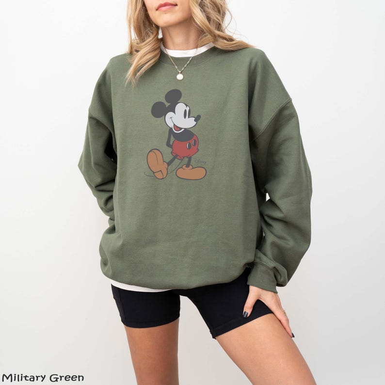 May include: An olive green sweatshirt featuring a classic Mickey Mouse graphic. The cartoon character is depicted in black, red, and tan colors. The sweatshirt is paired with black shorts. The text "Military Green" is at the bottom left.